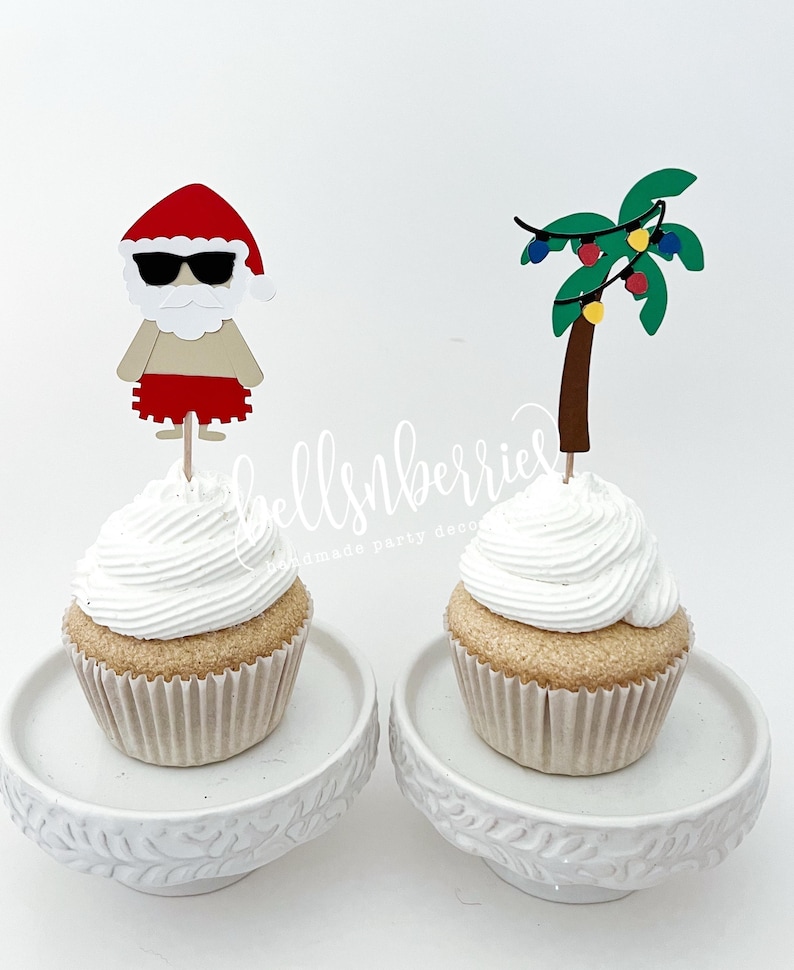 May include: Two cupcakes with white frosting, each on a white decorative plate. One cupcake has a Santa Claus topper with sunglasses, and the other has a palm tree with Christmas lights topper. The cupcakes are in paper liners.