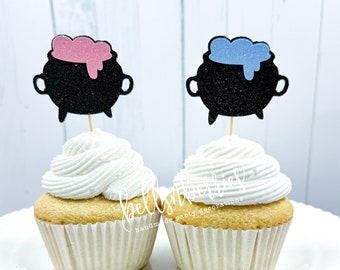 May include: Two cupcakes with black glitter cauldron cupcake toppers. One topper has pink frosting and the other has blue frosting. The cupcakes are frosted with white frosting.