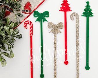 May include: Set of six Christmas-themed drink stirrers. The stirrers are made of acrylic and feature a variety of designs, including a candy cane, a bow, and a Christmas tree. The stirrers are red, green, and gold.