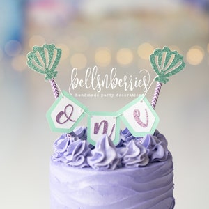 May include: Purple frosted cake with a green and purple "One" banner and two green glitter seashell toppers. The cake is decorated with purple frosting swirls. The banner reads "bellsnberries handmade party decorations".