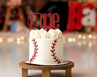 May include: A white frosted cake with red baseball stitching and a red glitter "one" cake topper. The cake sits on a small wooden stand. The cake is decorated with white frosting and is likely for a first birthday celebration.