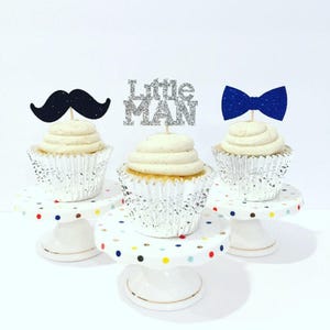 May include: Three cupcakes decorated with silver glitter cupcake toppers. The toppers are a black mustache, a silver glitter "Little Man" sign, and a blue bow tie. The cupcakes are on white cake stands with colorful polka dots.