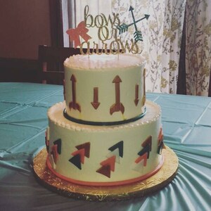 May include: A two-tiered white cake decorated with gold arrows and pink and blue bows. The cake is topped with a gold glitter sign that reads "bows or arrows".