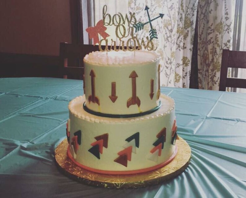 May include: A two-tiered white cake decorated with gold arrows and pink and blue bows. The cake is topped with a gold glitter sign that reads "bows or arrows".