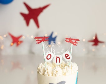 May include: A white cake with red and white frosting and a red airplane topper. The cake is decorated with a banner that says "one".