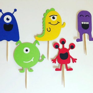 May include: Five colorful monster cupcake toppers made of felt. The toppers are in the shapes of a purple monster with a wide smile, a green monster with one eye, a red monster with three eyes, a yellow monster with one eye and a blue monster with one eye.