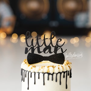 May include: A white cake with black chocolate drip icing and gold sprinkles. The cake is topped with a black glitter cake topper that says "little man" with a bow tie.