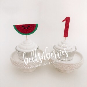 May include: Two cupcakes decorated with red glitter watermelon and red glitter number one toppers. The cupcakes are on a white cake stand with the text "bellsnberries handmade party decorations".
