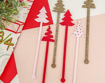 May include: A collection of Christmas tree-shaped drink stirrers in various colors. The stirrers include pink, red, and gold glitter designs. The stirrers are arranged on a pink and white background, with a red triangle and a festive patterned cloth in the background.