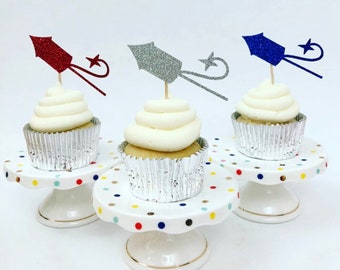 Könnte beinhalten: Drei Cupcakes, die mit Glitzer-Feuerwerkskörpern in Rot, Silber und Blau verziert sind. Die Cupcakes stehen auf weißen Tortenständern mit bunten Punkten.
