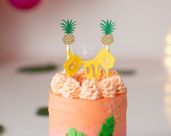 May include: A pink and orange ombre cake with a pineapple and "One" banner topper. The cake is decorated with green leaves and gold glitter.