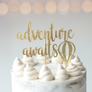 May include: A white cake with gold glitter frosting and a gold glitter topper that says "adventure awaits" with a hot air balloon design.