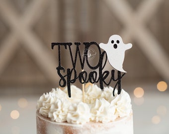May include: Black and white cake topper with the text "TWO spooky" and a white ghost. The topper is on a white frosted cake.