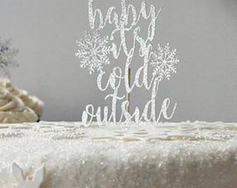 May include: Silver glitter cake topper with the text "Baby it's cold outside". The topper is on a white cake with white snowflake decorations.