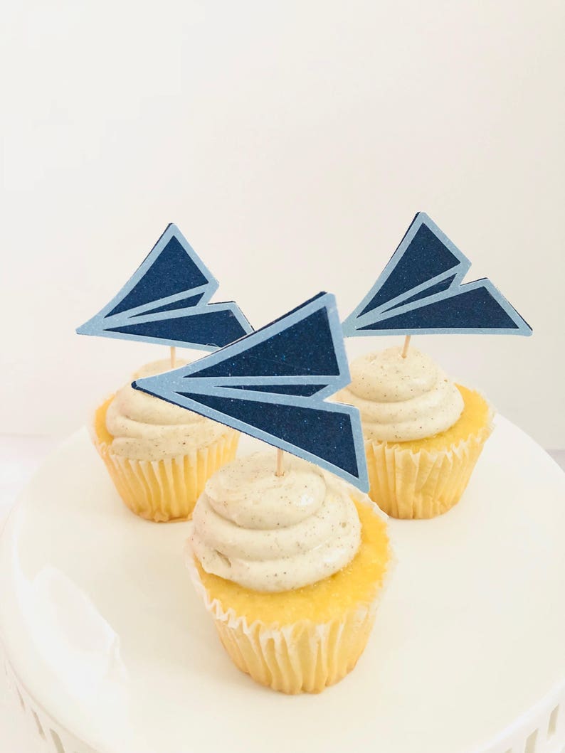 May include: Three cupcakes with vanilla frosting and blue paper airplane toppers. The cupcakes are on a white cake stand.