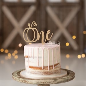 May include: A pink and white cake with a gold glitter pumpkin and the word "one" on top. The cake is on a wooden cake stand.