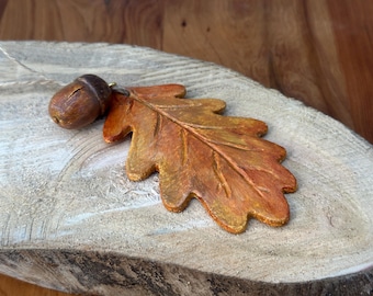 May include: A brown acorn charm hangs from a string with a golden brown oak leaf charm. The charms are on a wooden surface.