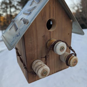 May include: Handmade wooden birdhouse with a license plate roof. The birdhouse features a round entry hole and is adorned with ceramic insulators and wire accents. The natural wood tones contrast with the metallic roof and insulators, creating a rustic aesthetic.