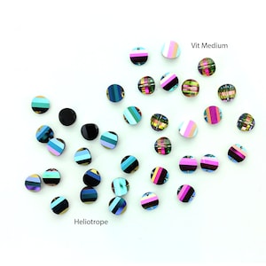 May include: Assortment of round, iridescent glass beads in various colors, including black, pink, blue, green, and white. The beads have a striped pattern and are arranged on a white surface. The text "Vit Medium" and "Heliotrope" are visible in the image.