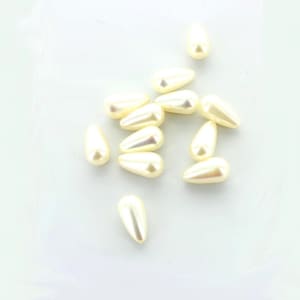 May include: A group of off-white pearlescent teardrop-shaped beads. The beads are scattered on a white surface.