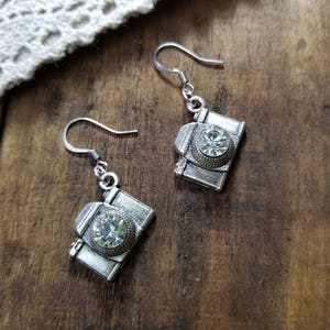 Puede incluir: Un par de pendientes de plata en forma de cámara con detalles de cristal de imitación transparente.
