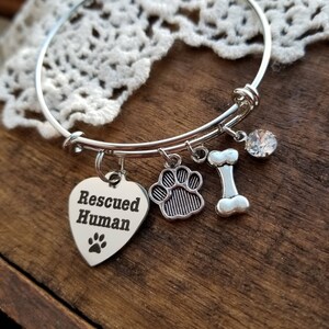 Puede incluir: Pulsera de plata con un dije en forma de coraz&oacute;n que dice "Rescued Human", un dije de huella de pata, un dije de hueso y un dije de cristal transparente.