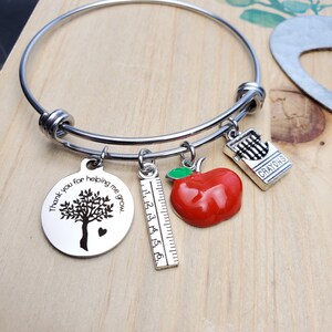 Könnte beinhalten: Ein silbernes Armband mit vier Anhängern: ein Baum mit dem Text "Danke, dass du mir beim Wachsen hilfst", ein roter Apfel, ein Lineal und eine Schachtel mit dem Text "Crayons".