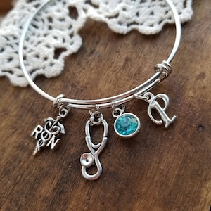 Könnte beinhalten: Ein silbernes Armband mit einem Stethoskop, einem blauen Kristall, einem RN-Anhänger und einem Buchstaben P-Anhänger.