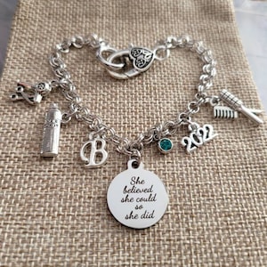 May include: A silver charm bracelet with a heart-shaped clasp and various charms, including a hair dryer, scissors, a bottle of hairspray, a letter "B", a small turquoise gem, the year "2022", and a round charm with the text "She believed she could so she did".