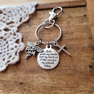 May include: Silver keychain with a medical symbol, a cross, and a round charm with the text "Lord, guide my hands and my heart as I help care for my patients today."