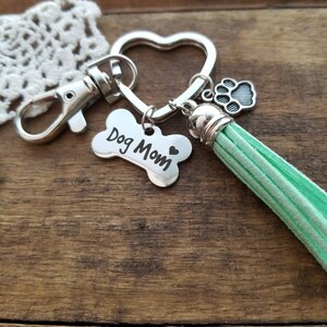 May include: Silver key chain with a heart-shaped ring, a bone charm with the text "Dog Mom" and a small paw print charm. The key chain also has a green tassel.