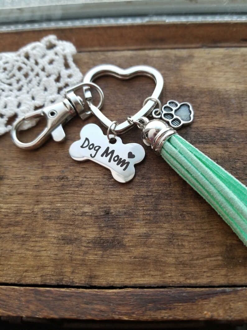 May include: Silver key chain with a heart-shaped ring, a bone charm with the text "Dog Mom" and a small paw print charm. The key chain also has a green tassel.