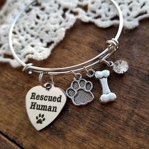 Puede incluir: Pulsera de plata con un dije en forma de coraz&oacute;n que dice "Rescued Human" y una huella de pata. La pulsera tambi&eacute;n tiene un dije de huella de pata, un dije de hueso y un dije de cristal transparente.