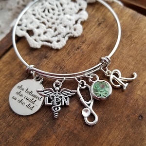 May include: A silver charm bracelet with a green gemstone, a stethoscope charm, a charm with the words "she believed she could... so she did", and a charm with the words "LPN" and a caduceus symbol.