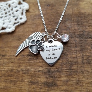 Puede incluir: Collar plateado con un colgante de corazón grabado con las palabras "a piece of my heart is in heaven". El collar también presenta un dije de huella de pata, un dije de ala y un cristal transparente. El collar se exhibe sobre una superficie de madera.