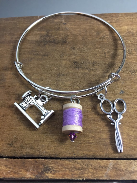 A silver charm bracelet on a wooden table. The charms are an antique sewing machine, a purple thread spool, and scissors.