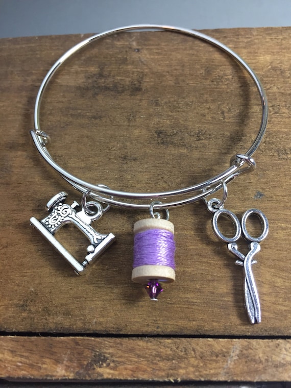 A silver charm bracelet on a wooden table. The charms are an antique sewing machine, a purple thread spool, and scissors. 