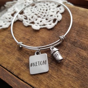 Op de afbeelding: Zilveren uitbreidbare armband met een vierkante bedel met de tekst "#KETOAF" en een kleine bedel in de vorm van een koffiekopje.