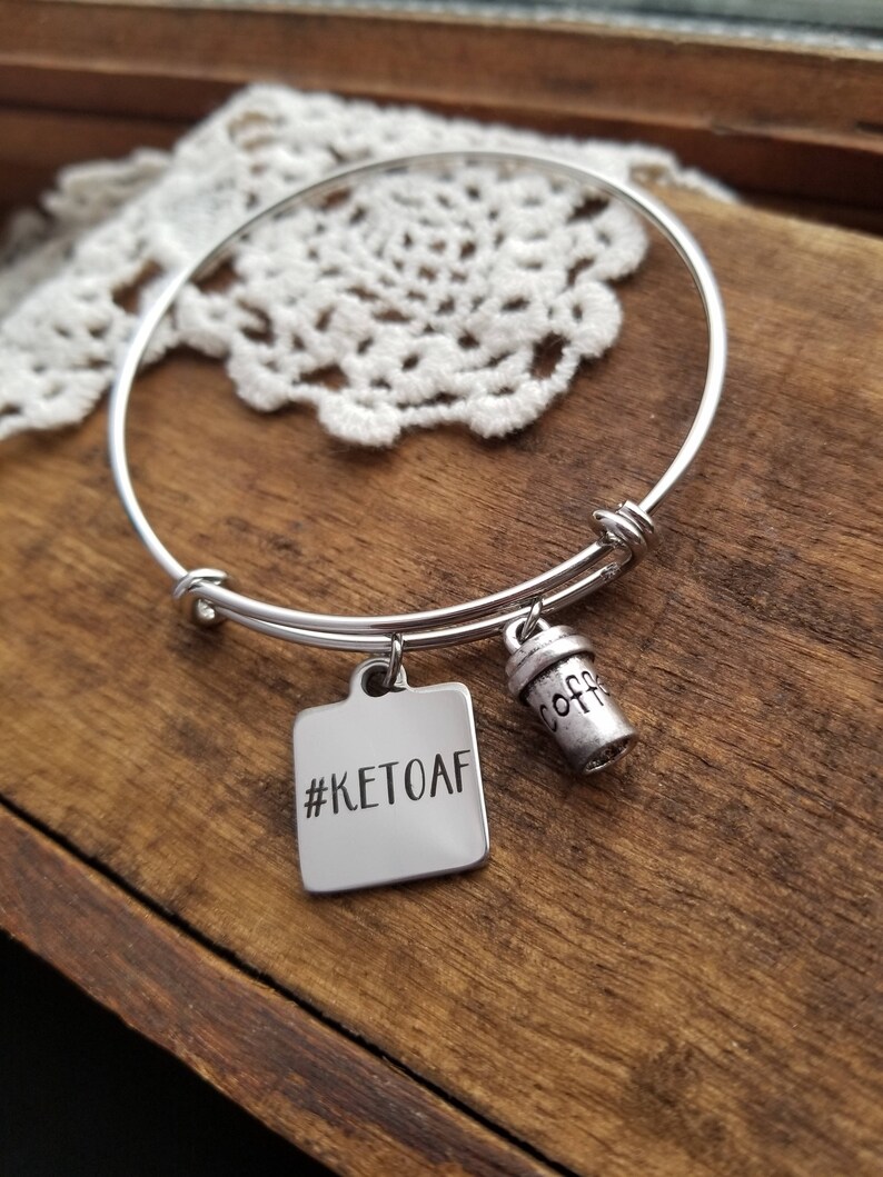 Op de afbeelding: Zilveren uitbreidbare armband met een vierkante bedel met de tekst "#KETOAF" en een kleine bedel in de vorm van een koffiekopje.
