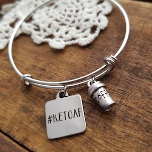 Op de afbeelding: Zilveren armband met twee bedels. Een bedel is een vierkant met de tekst "#KETOAF" en de andere is een koffiebekerbedel.