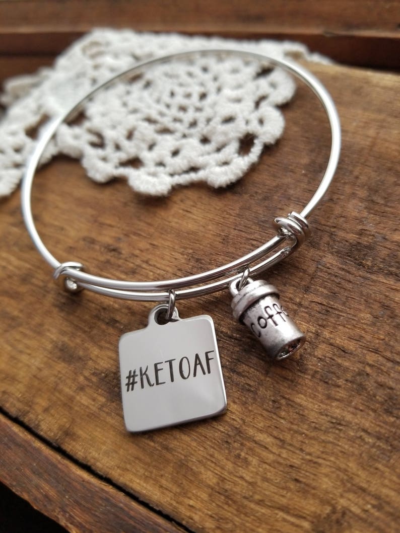 Op de afbeelding: Zilveren armband met twee bedels. Een bedel is een vierkant met de tekst "#KETOAF" en de andere is een koffiebekerbedel.