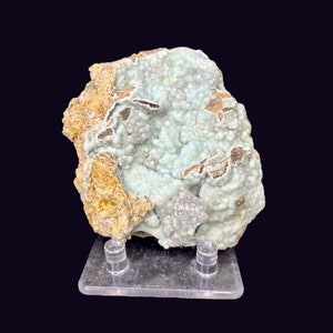 Puede incluir: Una roca grande de forma irregular con una formación mineral azul claro y blanco. El mineral tiene una textura burbujeante y está parcialmente cubierto por una matriz rocosa marrón. La roca se muestra en un soporte de acrílico transparente.