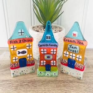 Puede incluir: Tres figuras de casas de cristal coloridas con los textos "Fish & Chips", "Cornish Candy" y "Cream Tea". Las casas tienen fachadas azules, naranjas y amarillas con detalles decorativos, incluyendo ventanas, puertas y pequeños pájaros. Una pequeña planta está en el fondo.
