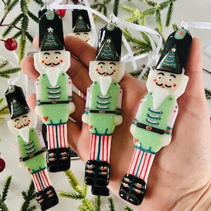 Könnte beinhalten: Vier grüne und weiße gläserne Nussknacker-Anhänger mit schwarzen Hüten und rot-weiß gestreiften Beinen. Jeder Anhänger hat einen kleinen grünen Weihnachtsbaum auf dem Hut.