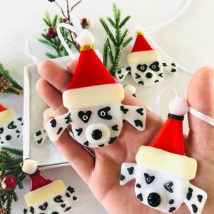 May include: Four handmade glass ornaments shaped like Dalmatian dog heads wearing Santa hats. The ornaments are white with black spots and have black eyes and noses. They are hanging from white ribbon.