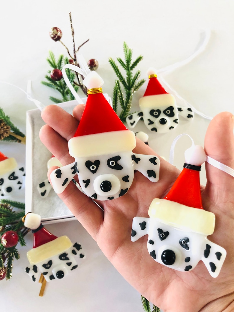 May include: Four handmade glass ornaments shaped like Dalmatian dog heads wearing Santa hats. The ornaments are white with black spots and have black eyes and noses. They are hanging from white ribbon.