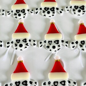 May include: Nine handmade glass ornaments shaped like Dalmatian dogs wearing Santa hats. The ornaments are white with black spots and have black eyes and noses. Each ornament has a white ribbon attached for hanging.
