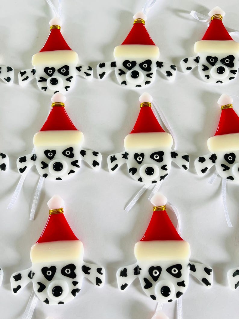 May include: Nine handmade glass ornaments shaped like Dalmatian dogs wearing Santa hats. The ornaments are white with black spots and have black eyes and noses. Each ornament has a white ribbon attached for hanging.