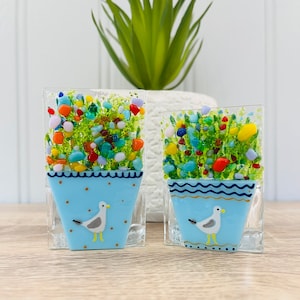 May include: Two small glass mosaic flower pots with blue and white designs. Each pot features a seagull and a colourful flower arrangement.