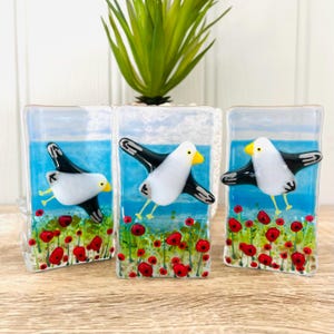 Könnte beinhalten: Drei Glas-Kunstwerke, die fliegende Möwen über einem Feld roter Mohnblumen darstellen. Jedes Stück zeigt eine weiße Möwe mit schwarzen Flügeln vor einem blauen Himmel und Meereshintergrund. Die Mohnblumen sind rot mit grünen Stielen.